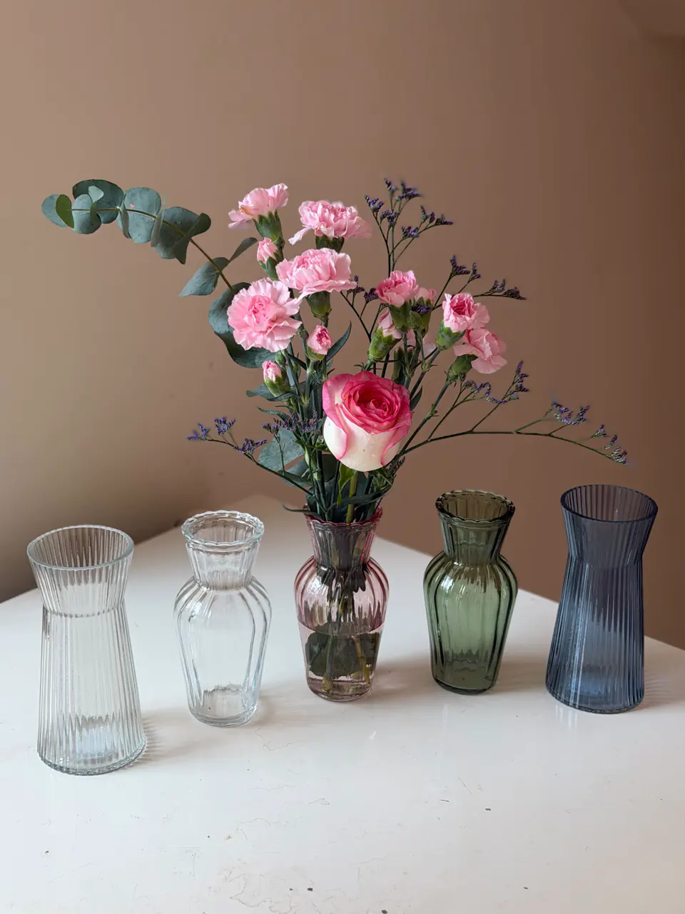 Arranjo floral com cravos rosa e eucalipto em vaso de vidro, ao lado de vasos decorativos