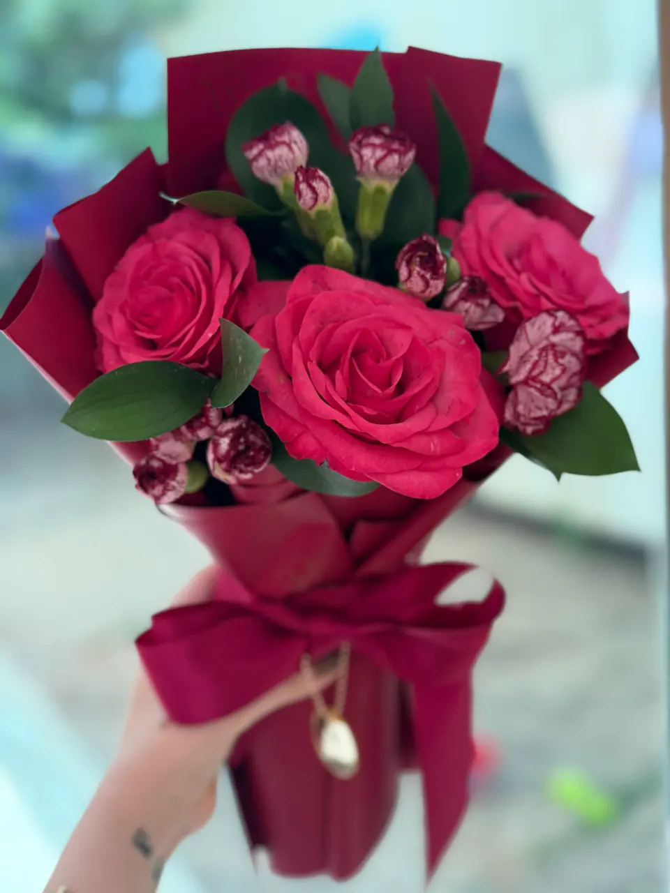 Mãos entregando buquê de rosas pink com cravos e rusco em papel vinho